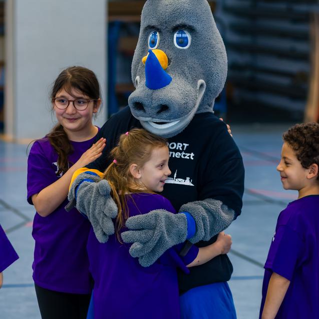 Maskottchen umarmt Kinder in lila T-Shirts während einer Sportveranstaltung in einer Turnhalle.