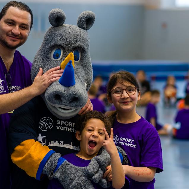 Gruppenselfie mit einem Maskottchen und zwei Kindern in lila T-Shirts, umgeben von weiteren Kindern im Hintergrund.