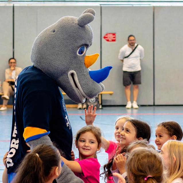 Ein Maskottchen interagiert mit mehreren Kindern in pinken T-Shirts in einer Sporthalle.