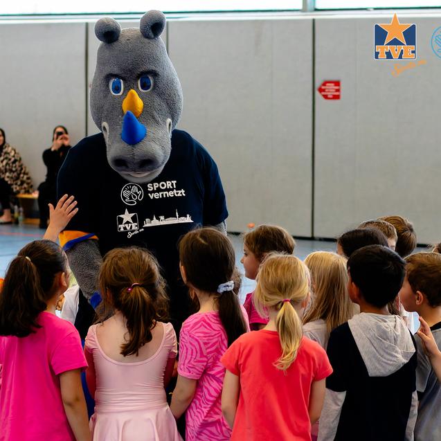 Ein Maskottchen in einem Sportshirt steht im Mittelpunkt, umringt von fröhlichen, bunten Kindern in einer Halle.