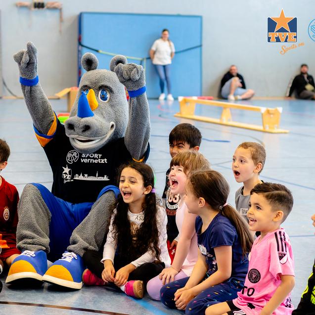 Gruppe von Kindern sitzt lachend um ein Maskottchen herum, in einer Sporthalle bei einem fröhlichen Event.