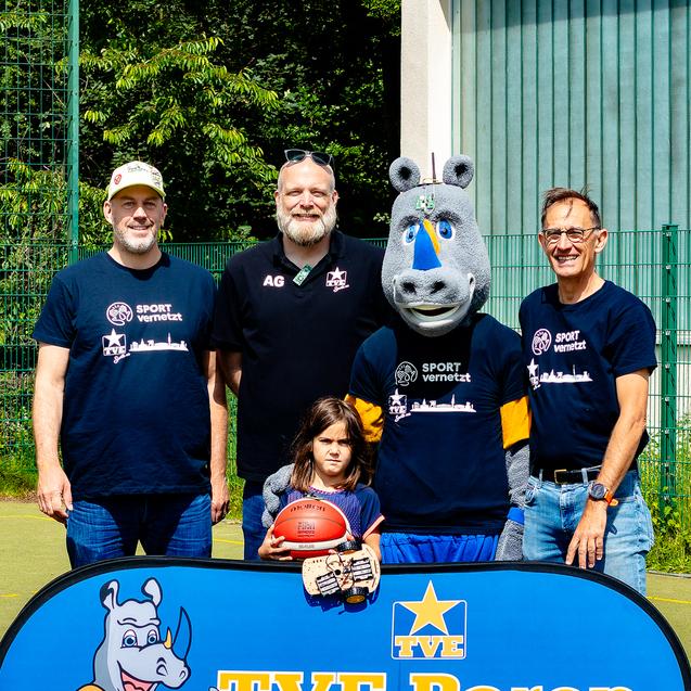 Gruppe von fünf Personen, darunter ein Maskottchen, posiert an einem Sportplatz vor einem Banner der TVE Barop.