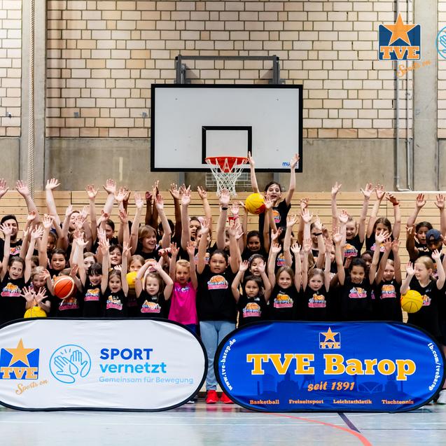 Gruppendynamik von Kindern und Trainern in Sportkleidung, die mit Basketballbällen auf einem Hallenboden posieren.