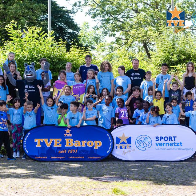 Gruppenfoto von Kindern und Trainern in blauen T-Shirts vor einer grünen Kulisse, begleitet von zwei Banner.