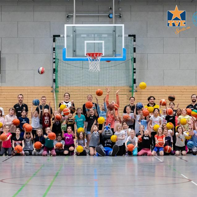 Gruppenshooting von Kindern und Betreuern in Sportkleidung, die Basketballbälle in die Luft werfen, in einer Halle.