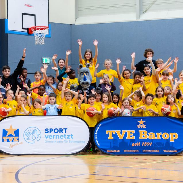Gruppensfoto von Kindern und Trainern in gelben T-Shirts, die im Basketball-Hallenraum jubeln. Banner im Vordergrund.