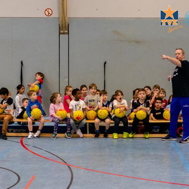 Gruppe von Kindern sitzt auf einer Bank in einer Sporthalle und hält Basketballs, während ein Trainer mit ihnen spricht.