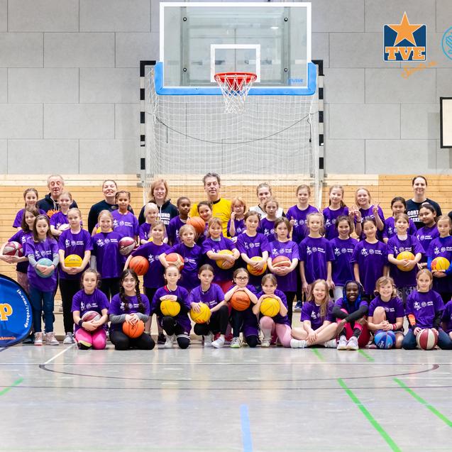 Gruppe von Mädchen in lila T-Shirts hält Basketballs, posiert auf einem Sportplatz vor einem Basketballkorb und Banner.