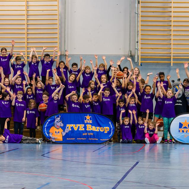 Gruppenfoto von Kindern in lila T-Shirts, die jubelnd in einer Sporthalle stehen, umgeben von Werbebannern.
