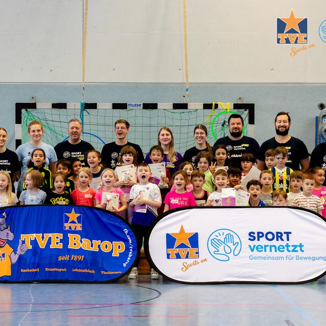 Gruppenfoto von Kindern und Trainern in einer Sporthalle, mit Bannern der Organisationen TVE Barop und SPORT vernetzt.