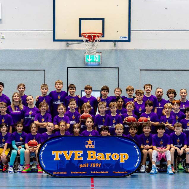 Gruppenshot von Kindern in lila Trikots vor einem Basketballkorb, mit einem Banner „TVE Barop“ im Vordergrund.
