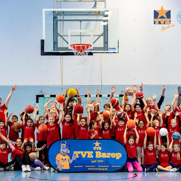 Gruppe von Kindern und Trainern in roten T-Shirts, die auf einem Basketballfeld mit Bällen und einem Banner posieren.
