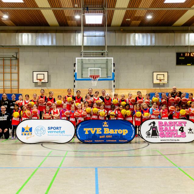Gruppenfoto einer Basketballmannschaft mit Spielern, Trainern und zwei Banner in einer Sporthalle.