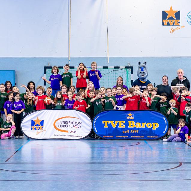 Gruppenfoto von Kindern und Erwachsenen in Sportkleidung in einer Turnhalle mit Basketball und Banner der Organisationen.