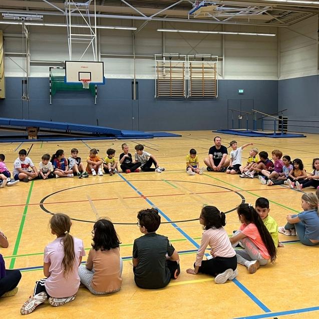 Gruppe von Kindern sitzt im Kreis auf dem Boden in einer Sporthalle, während Betreuer in der Nähe stehen.