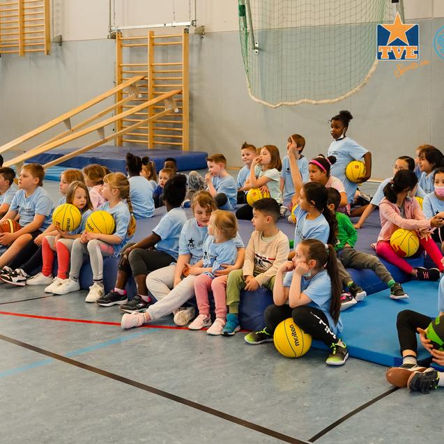 Gruppierung von Kindern in blauen T-Shirts, die auf Matten sitzen und Basketballbälle halten, in einer Sporthalle.