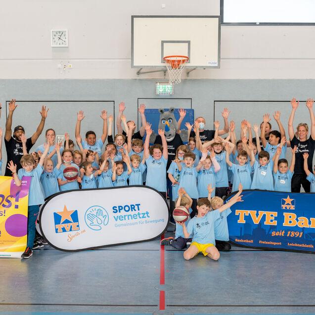 Gruppenfoto von Kindern und Trainern in Basketballshirts, die in einer Halle mit erhobenen Händen posieren.