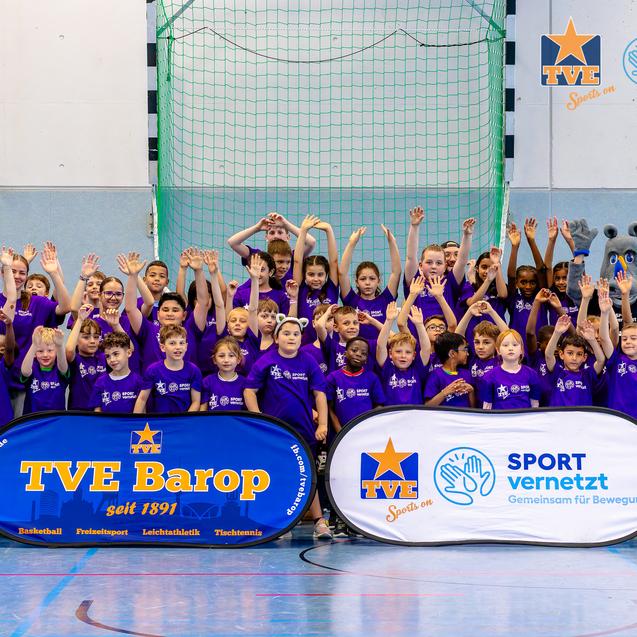 Gruppenerfoto von Kindern und Betreuern in lila T-Shirts, die in einer Sporthalle mit zwei Werbebannern posieren.
