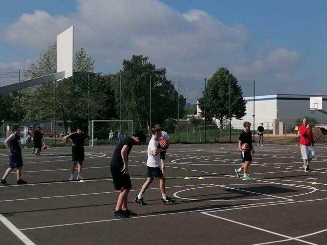 Gruppe von Jugendlichen spielt Basketball auf einem Freiplatz mit Basketballkörben unter blauem Himmel.