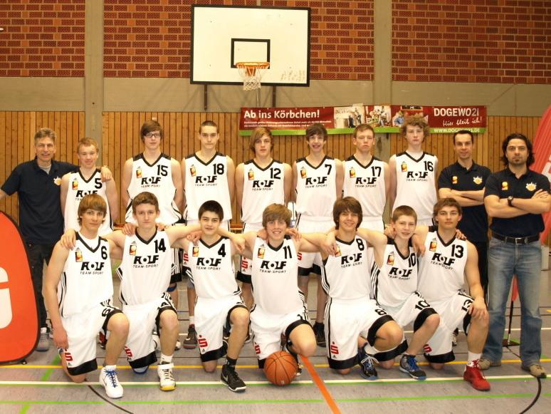 Juniorbaskets Gruppe von 15 Basketballspielern in weißen Trikots, posierend in einer Sporthalle mit Sponsorenlogos.