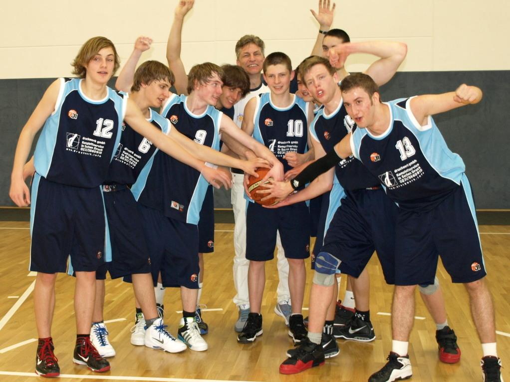 Gruppfoto von zehn Basketballspielern, die zusammen einen Ball in Richtung Kamera halten, im Sportumfeld.