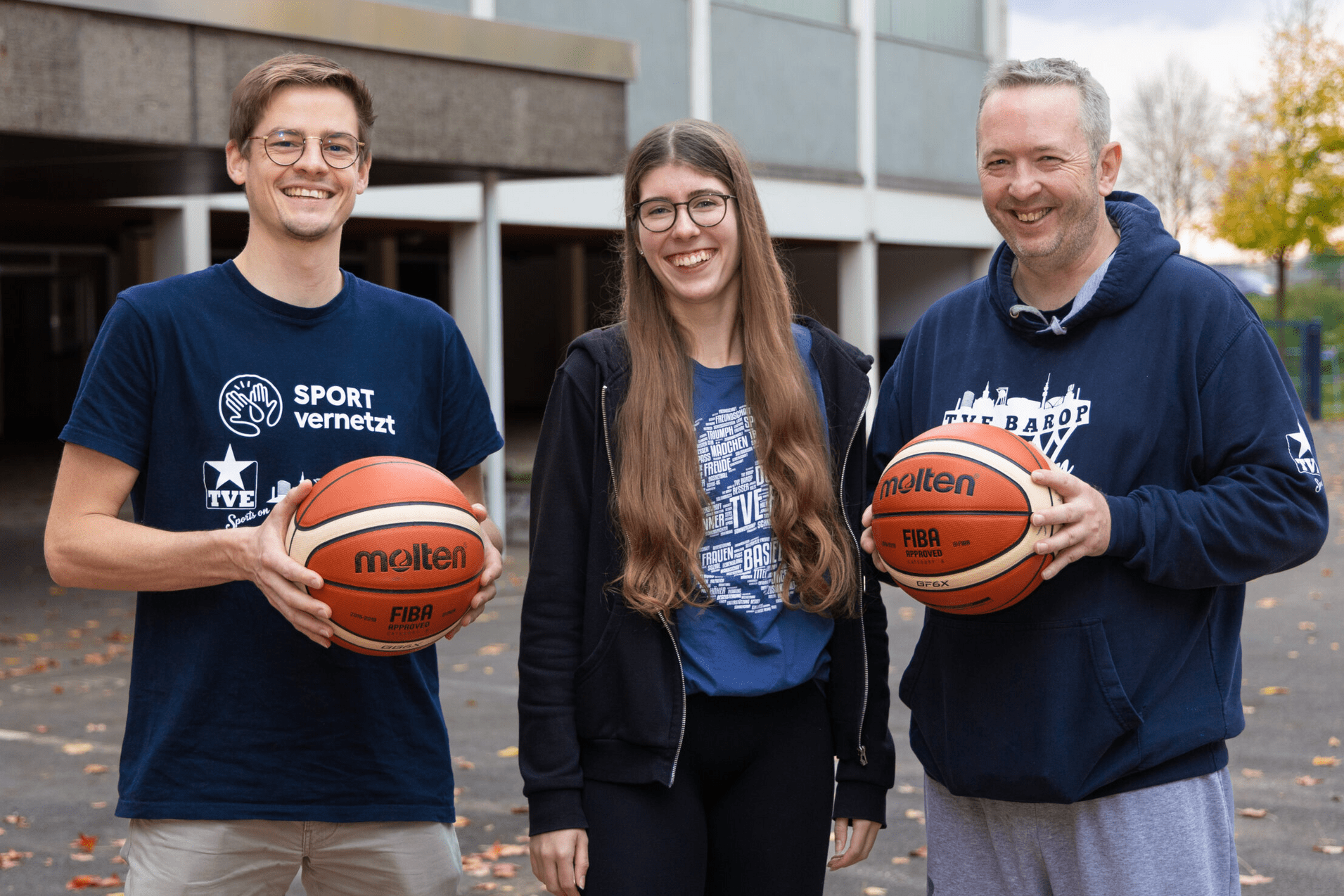 Hauptamtliche Drei Personen stehen lächelnd im Freien und halten Basketballs; im Hintergrund ist eine Sporteinrichtung sichtbar.