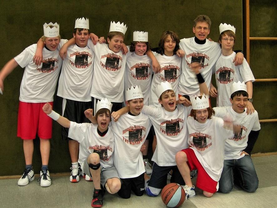 U14 2008 Gruppe von Kindern in weißen T-Shirts und Papierkrönen, lächelnd um einen Basketball versammelt.