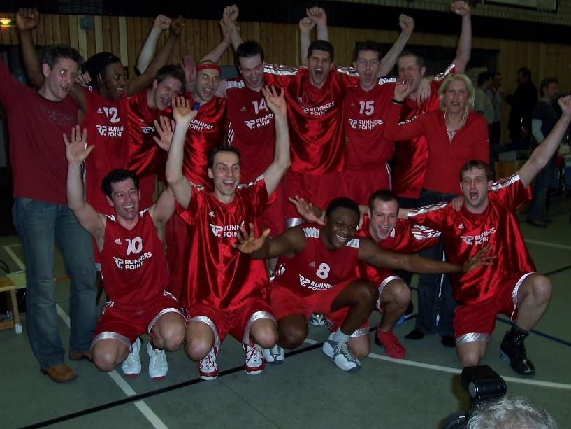 Herren 2003/04 Gruppenselfie einer Basketballmannschaft in roten Trikots, die jubelnd und freudig in einer Sporthalle posiert.