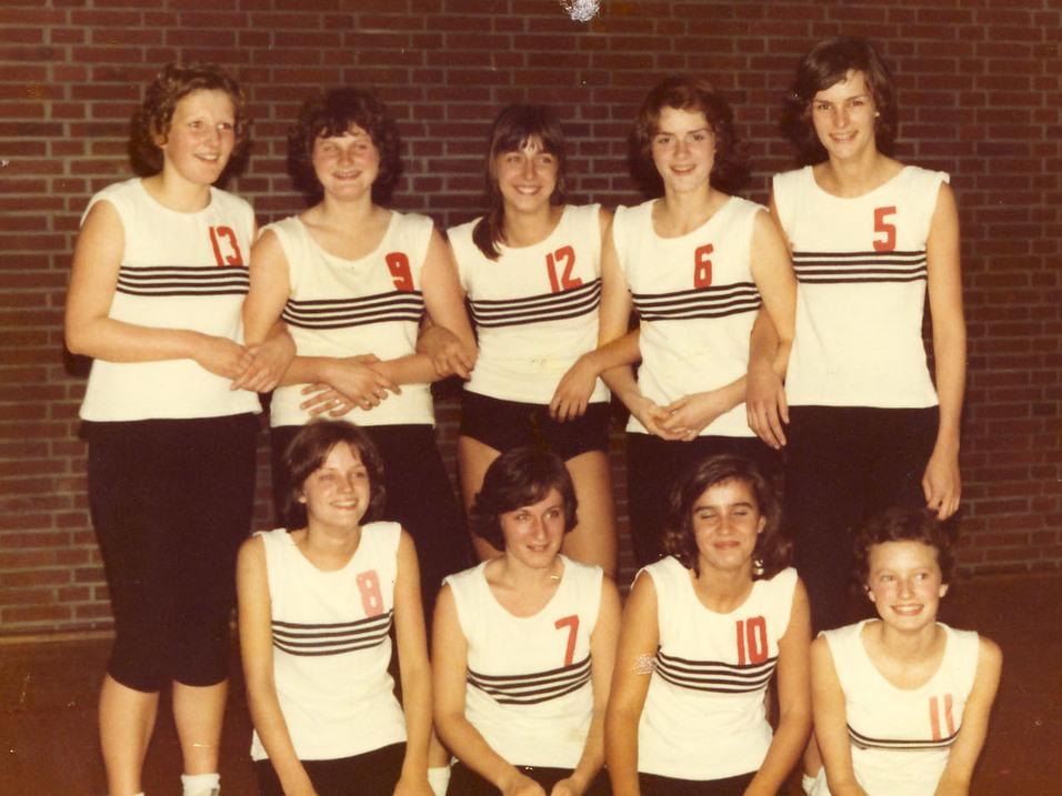 Mädchen 1973/74 Gruppenfoto von neun Frauen im Sportoutfit mit nummerierten Shirts in einer Sporthalle vor einer Ziegelwand.