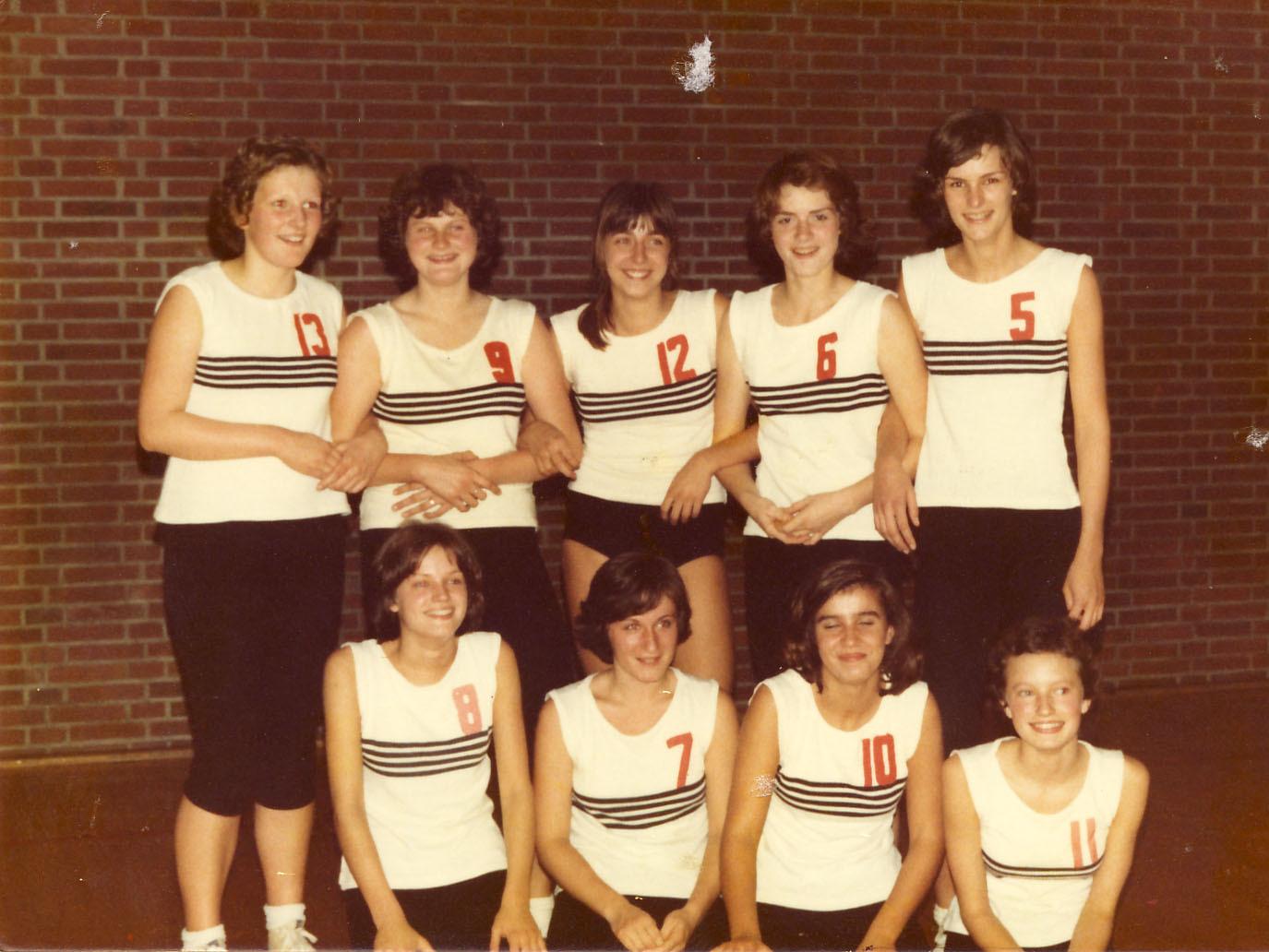 Mädchen 1973/74 Gruppenfoto von neun Frauen im Sportoutfit mit nummerierten Shirts in einer Sporthalle vor einer Ziegelwand.