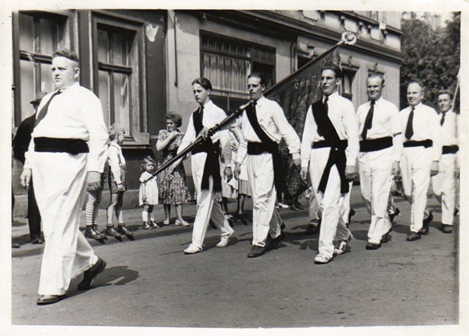 Männer in weißen Anzügen mit schwarzen Gürteln und Fahne ziehen in einer Parade durch eine Stadtstraße.