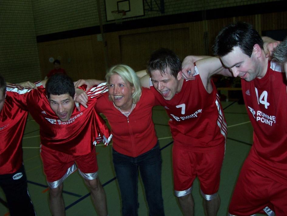 Alice Michalak Gruppe von Sportlern in roten Trikots umarmt eine Frau, alle lachen und feiern in einer Sporthalle.