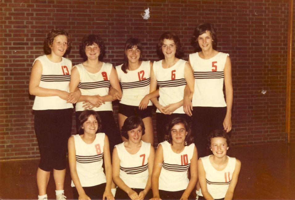 Mädchen 1973/74 Gruppenfoto von neun Frauen im Sportoutfit mit nummerierten Shirts in einer Sporthalle vor einer Ziegelwand.