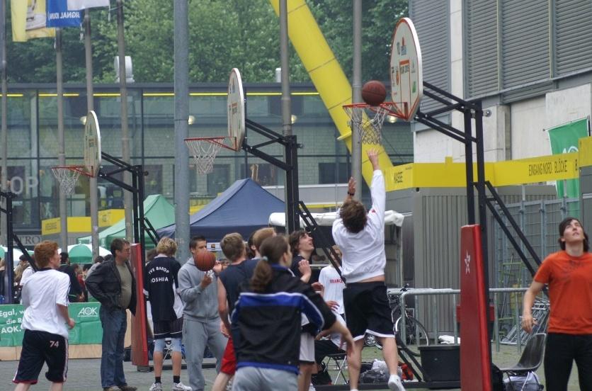 Jugendliche spielen Basketball auf einem Außenplatz, einige werfen den Ball zu den Körben.