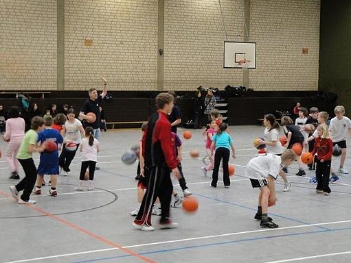 Gruppe von Kindern und Trainern beim Basketballtraining in einer Sporthalle, jeder mit einem Basketball in der Hand.