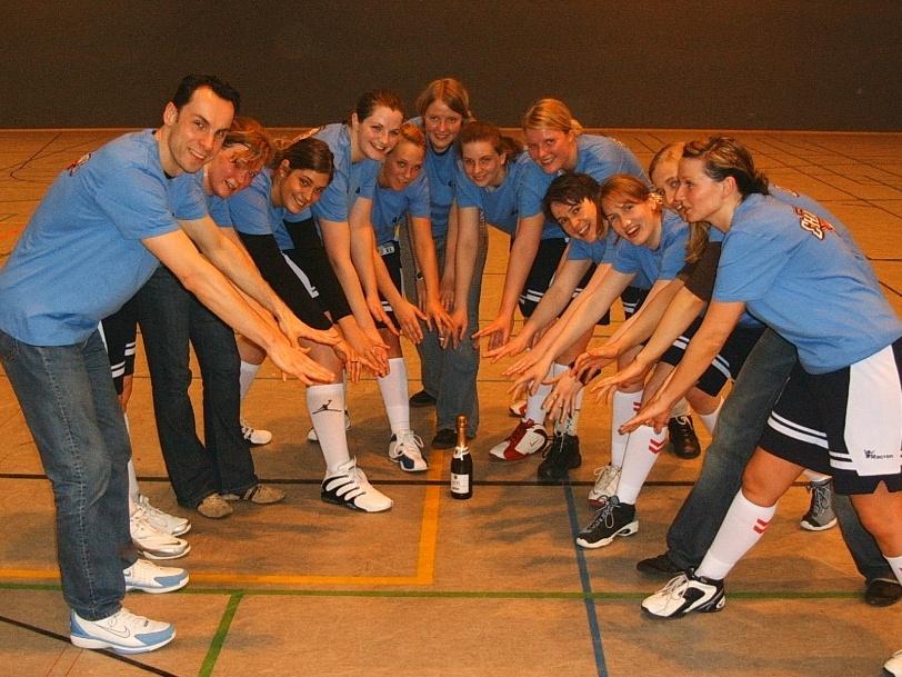 Damen 2006 Gruppe von 12 Personen in blauen T-Shirts im Hallensport, die um eine Flasche Sekt auf dem Boden posieren.