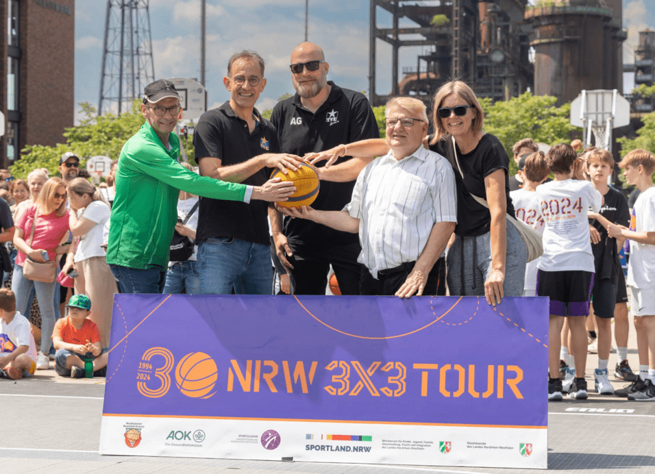 NRW Tour 2024 Fünf Personen halten gemeinsam einen Basketball vor einem Banner der NRW 3x3 Tour, mit Zuschauern im Hintergrund.