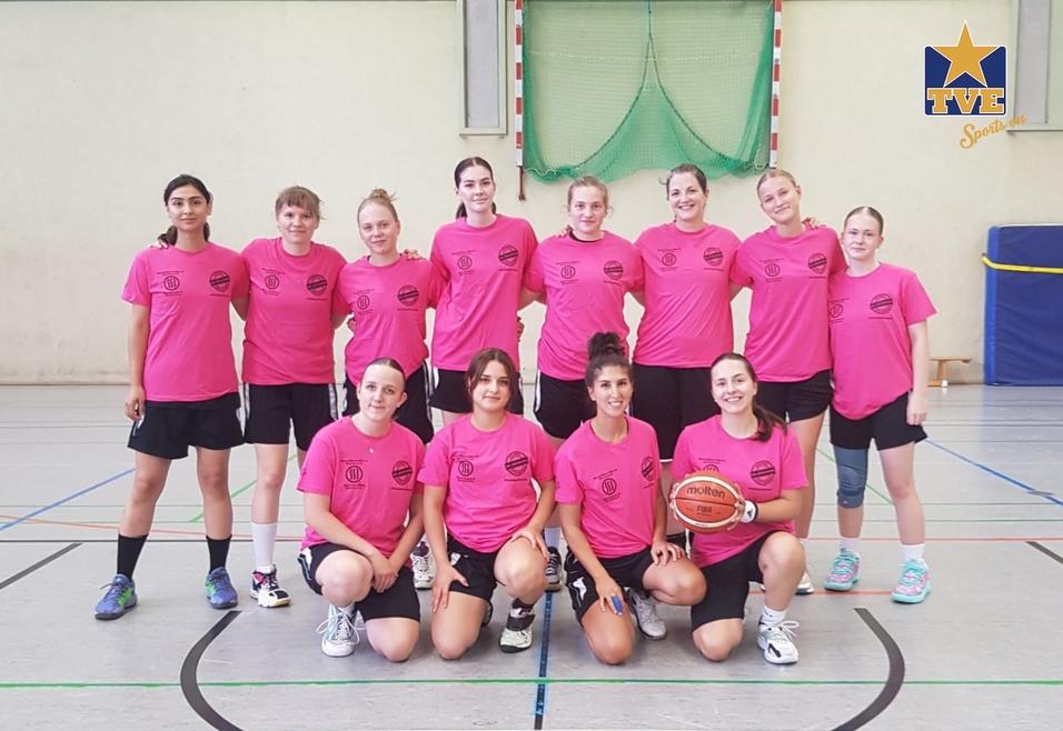 Gruppe von 12 Basketballspielerinnen in pinken Trikots, auf einer Sporthalle stehend, zusammen mit einem Basketball.