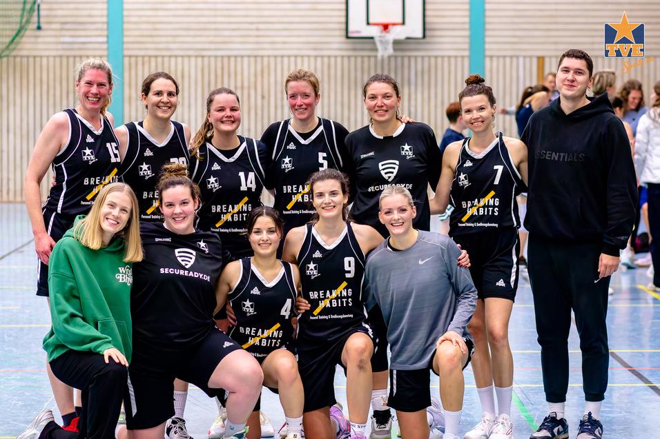 Gruppe von Basketballspielerinnen in einheitlichen Trikots posiert lächelnd auf dem Basketballfeld.