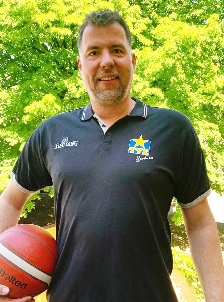 Mann in schwarzem Polo-Shirt hält einen Basketball, mit grünem Laub im Hintergrund.