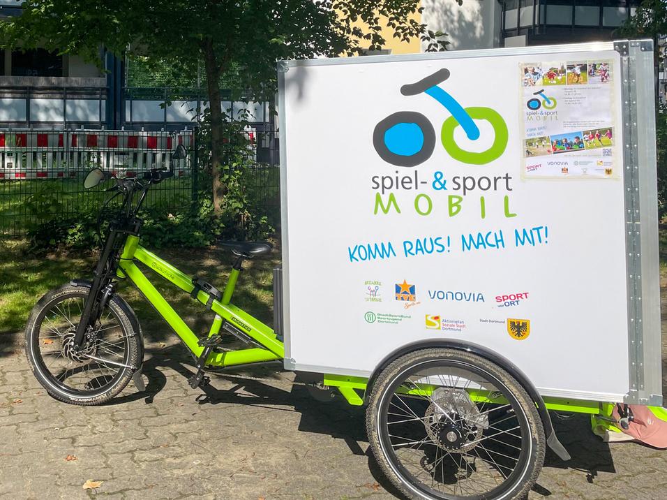 Fahrradanhänger mit Aufschrift „spiel- & sport MOBIL“ steht vor einem Wohngebäude unter Bäumen auf einem gepflasterten Weg.