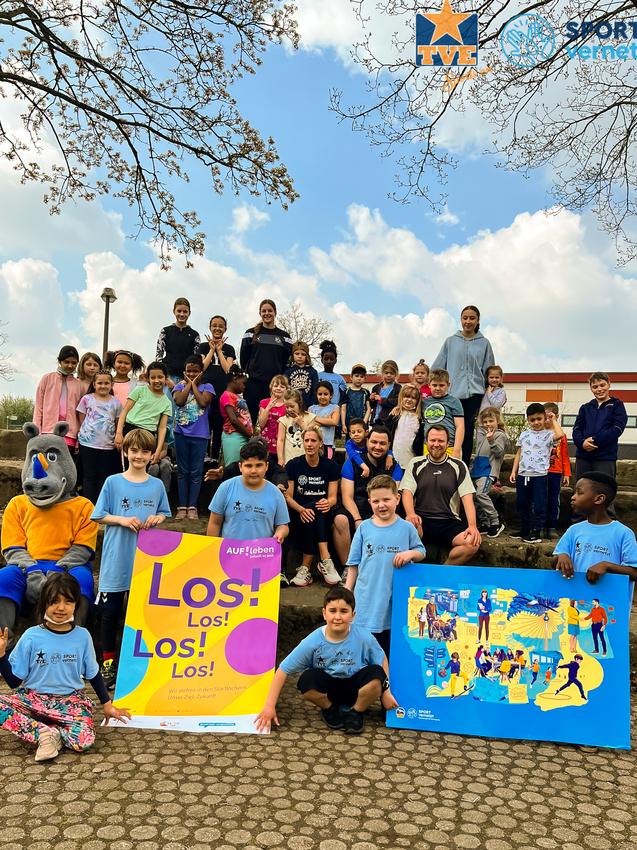 Gruppenselfie von Kindern und Betreuern in blauen T-Shirts, die mit bunten Plakaten im Freien posieren.