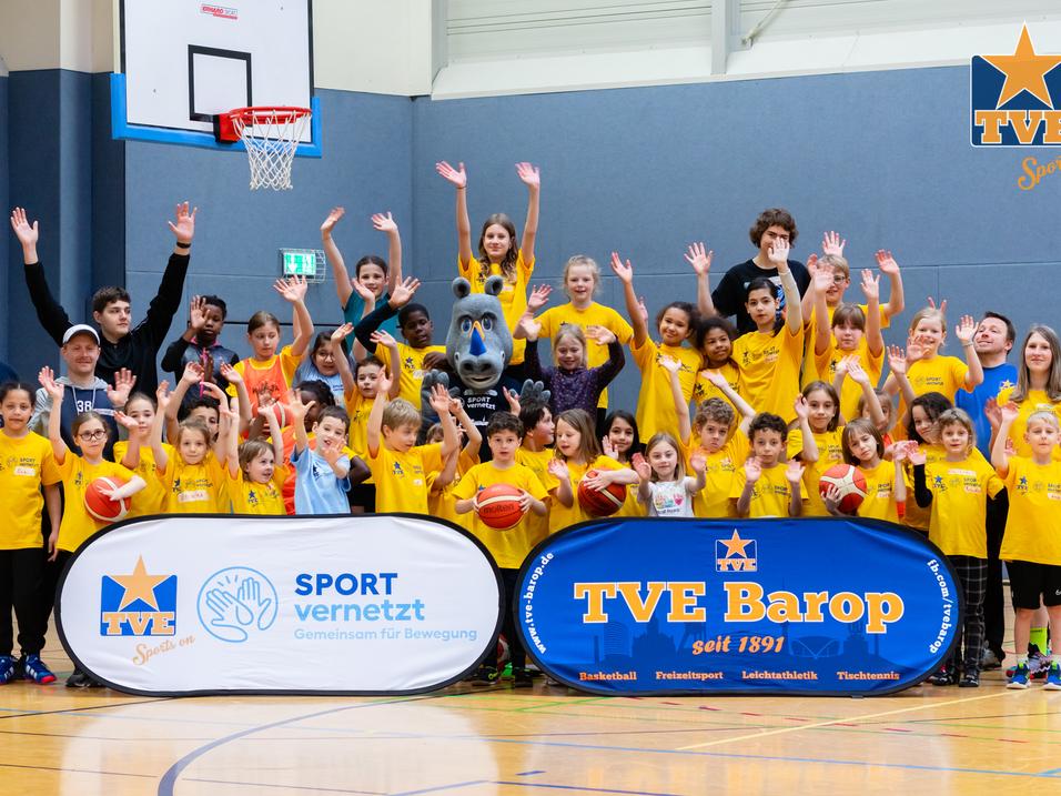 Gruppenshot von Kindern und Trainern in gelben T-Shirts, die mit Basketballs posieren, vor Sportbanner in einer Turnhalle.