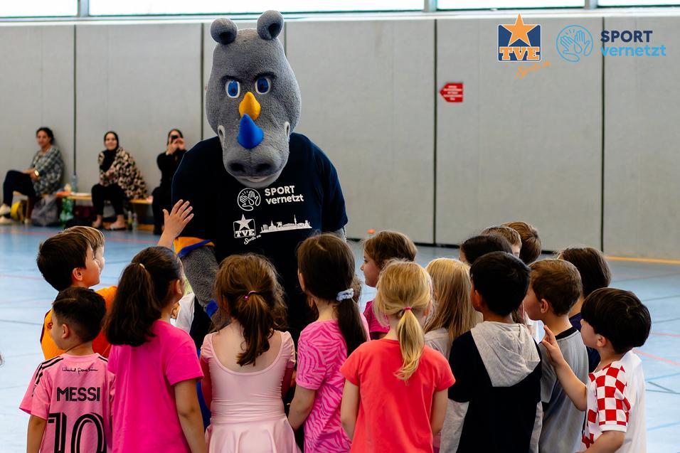 Rhingo Integration Ein Maskottchen in einem T-Shirt interagiert mit einer Gruppe von Kindern in einer Sporthalle während einer Veranstaltung.