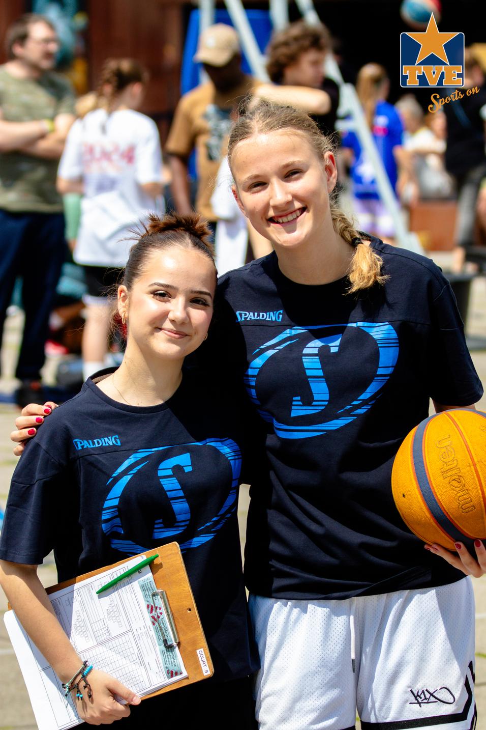 Zwei Mädchen in schwarzen T-Shirts stehen lächelnd zusammen, eines hält einen Basketball, das andere eine Clipboard.