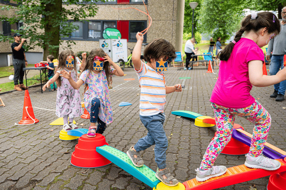 Kinder balancieren auf bunten Spielgeräten in einem Außenbereich, während Erwachsene im Hintergrund beobachten.