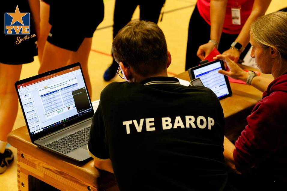 Zwei Personen analysieren Daten auf Laptops und Tablets während einer sportlichen Veranstaltung, im Hintergrund Sportler.
