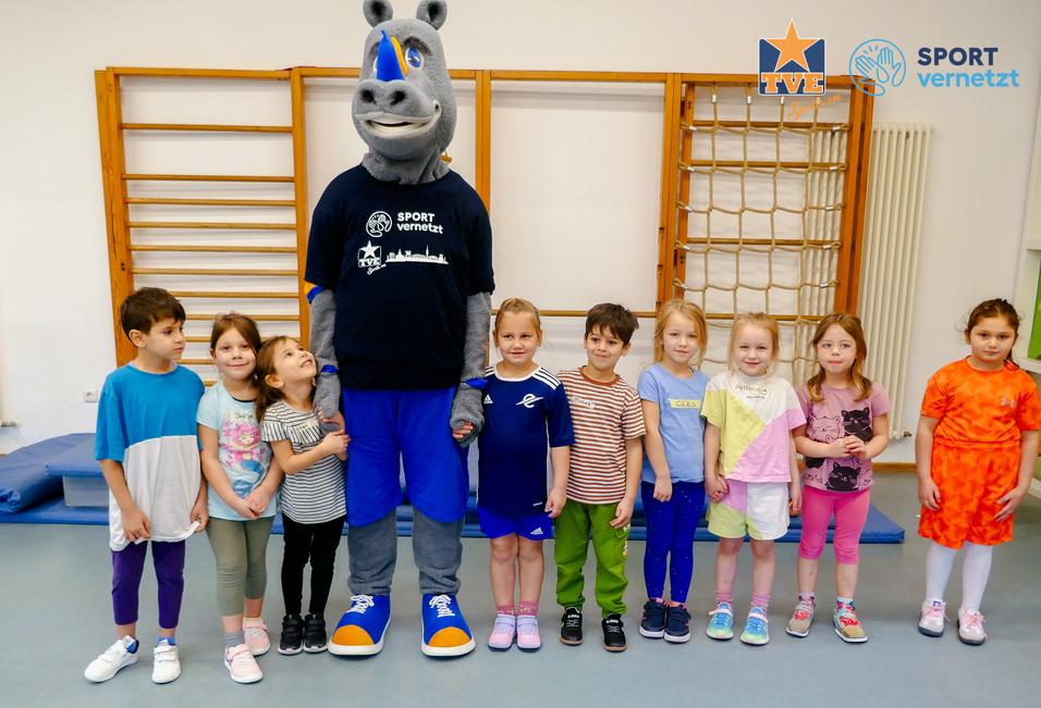 Rhingo Kita Gruppe von Kindern in Sportkleidung posiert mit einem Maskottchen in einem Turnraum.