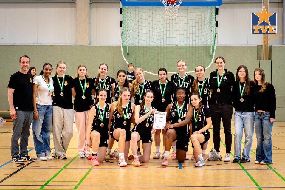 Man sieht ein weibliches Basketballteam in schwarzen Trikots mit Medaillen, posierend vor einem Basketballkorb.