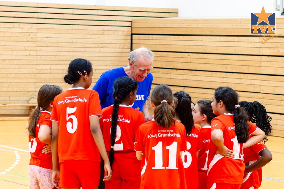 Trainer spricht motivierend mit einer Gruppe junger Mädchen im Basketballtrikot in einer Sporthalle.